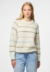 Mlada žena u kremastom pletenom svjetru s pastelnim horizontalnim trakama i svijetlo plavim trapericama, stoji naslonjena na običnu bijelu pozadinu.