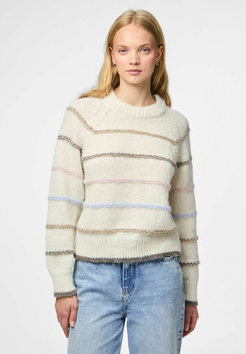 Mlada žena u kremastom pletenom svjetru s pastelnim horizontalnim trakama i svijetlo plavim trapericama, stoji naslonjena na običnu bijelu pozadinu.