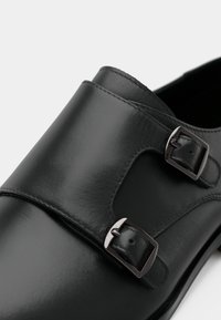 Μαύρα δερμάτινα παπούτσια που διαθέτουν δύο κούμπες κλεισίματος, λεία επιφάνεια και ραμμένες λεπτομέρειες στο σχέδιο του άνω μέρους.