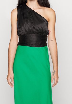 Mujer con una camiseta negra sin mangas y de un solo hombro con una cinturilla ancha y una falda verde brillante, mostrando tatuajes en ambos brazos.