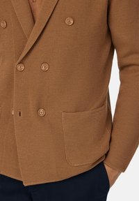 Blazer marrón texturizado con cierre de botones de doble botonadura, bolsillo frontal y sutil patrón de espiga. Combinado con pantalones azul marino.