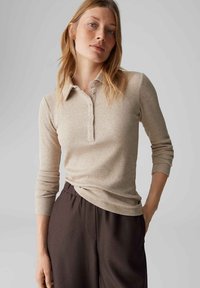Vrouw draagt een beige geribd poloshirt met lange mouwen en donkerbruine broek, staand met één hand in de zak tegen een effen grijze achtergrond.