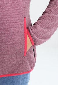 Pull polaire couleur lie de vin chiné avec des accents roses. Comprend une poche latérale et un ourlet contrastant. Manches longues avec tissu texturé.