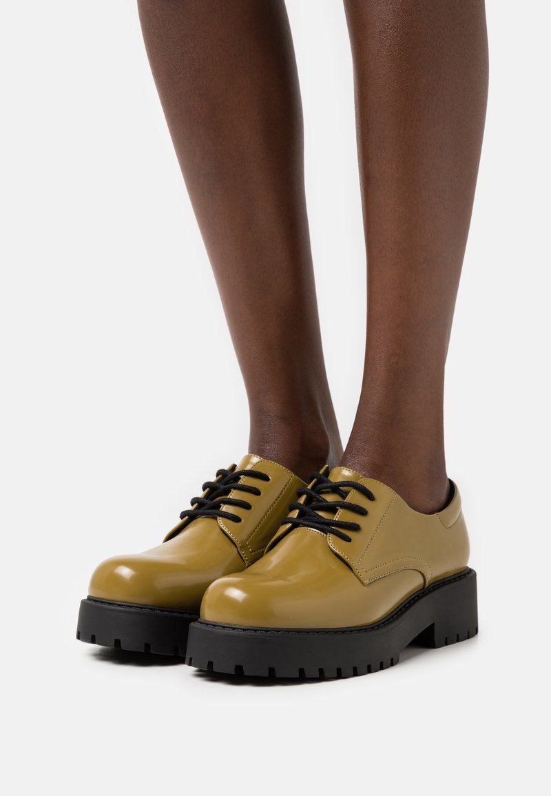 Chaussures à lacets vert olive brillantes avec semelles épaisses noires portées pieds nus sur fond blanc.