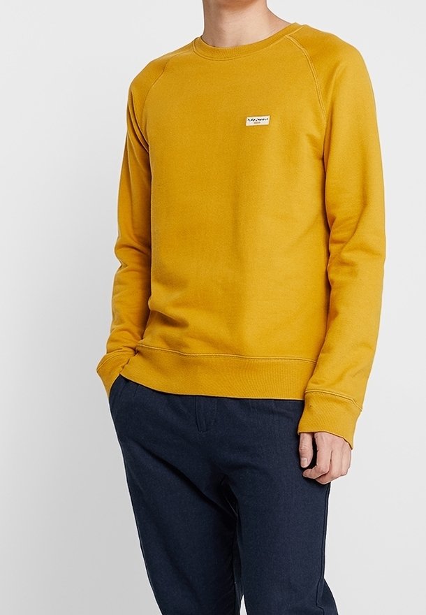 Homme portant un sweat-shirt à manches longues jaune moutarde avec un petit logo sur la poitrine, assorti à un pantalon bleu foncé, debout devant un fond uni.