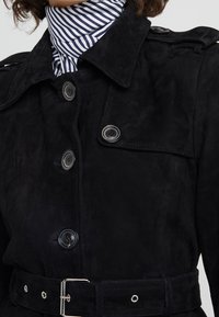 Vrouw in een zwarte suede trenchcoat met grote knopen, een riem en een zwart-wit gestreepte coltrui eronder.