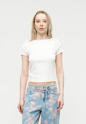 Mujer joven con cabello largo y rubio que lleva una blusa blanca de manga corta y jeans azules con patrones florales rosas y un cinturón atado.