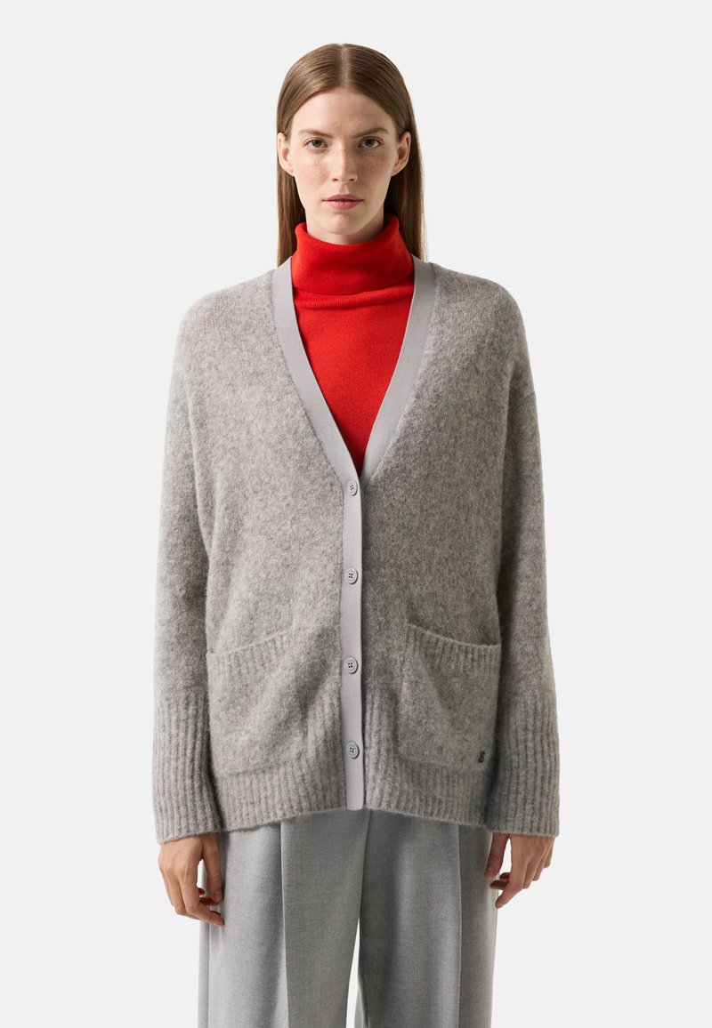 Grauer Wollcardigan mit V-Ausschnitt, hellgrauem Saum und zwei Vordertaschen. Verfügt über gerippte Bündchen. Wird über einem roten Rollkragenpullover getragen.