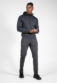 Grijze sportieve zip-up jas met capuchon en zakken, gedragen met bijpassende taps toelopende broek. Materiaal lijkt glad met een elegante designaccenten.