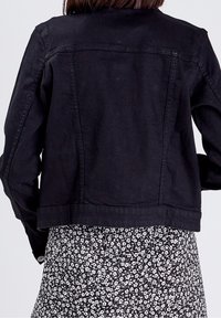 Zwarte spijkerjack met een getailleerd ontwerp, voorzien van een achterpand naad en een onafgewerkte zoom. Gedragen over een zwart-wit gebloemd patroonjurk.