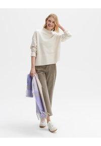 Cremfarbener Strickpullover mit hohem Kragen, kombiniert mit olivgrünen weit geschnittenen Hosen und einem gefransten Schal in Lila und Beige.