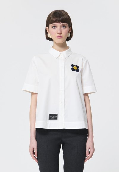 Chemise blanche à manches courtes avec col, ornée d'une broderie de fleurs bleu marine et d'une étiquette noire en bas. Tissu en coton lisse.