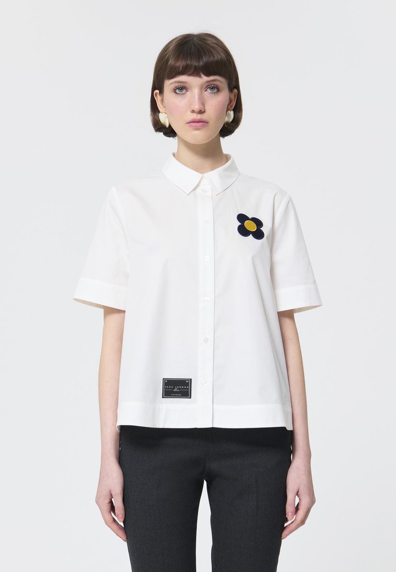 Witte kortarmige shirt met een kraag, voorzien van een marineblauwe bloemenborduring en een zwart label aan de zoom. Gladde katoenen stof.