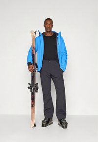 Smučarska oprema s svetlo modro jakno, črno majico, črnimi hlačami in črnimi smučarskimi čevlji, stoji na smučeh na belem ozadju.