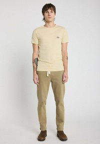T-shirt en coton beige avec un col rond et un petit motif graphique sur la poitrine, associé à un pantalon kaki et des bottines marron.