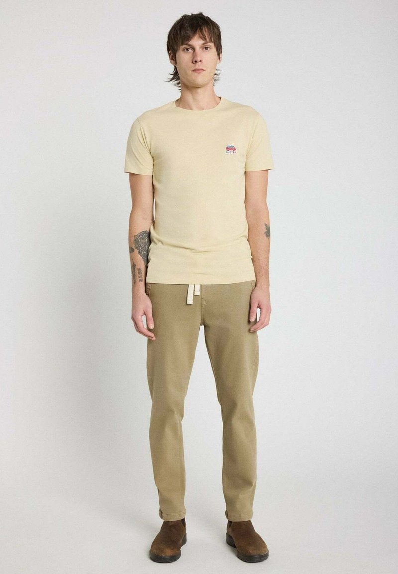 T-shirt en coton beige avec un col rond et un petit motif graphique sur la poitrine, associé à un pantalon kaki et des bottines marron.