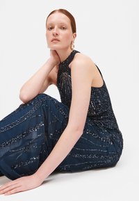 Abito blu navy con scollo all'americana, ornato da dettagli intricati di paillettes, caratterizzato da un corpetto aderente e una gonna fluttuante con motivi ondulati.