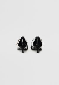 Musta patenttinaahkakengät, joissa on teräväkärkinen muotoilu ja pieni takavetoketjuyksityiskohta. Hoikka, kapea stiletto-kanta. Sujuva pinta.