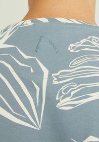 Vaaleansininen kangastoppi, jossa on valkoisia lehtikuvioita, näkyvillä ompeluyksityiskohdilla ja pyöreällä kaula-aukolla takana.