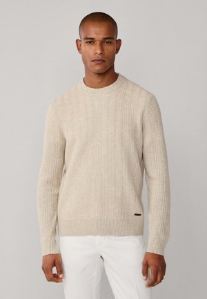 Beige gebreide trui met ronde hals, geribbelde manchetten en zoom, met een lichte structuur en een klein zwart embleem aan de onderkant links.