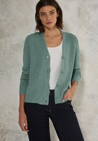 Ljust teal-färgad cardigan med en texturerad ribbad design, V-ringning, knappar fram och långa ärmar, i kombination med en vit topp och mörka jeans.