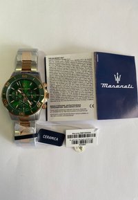 Maserati ur med grøn skive, guld- og sølvfarvet rustfrit stålarmbånd, chronograph-funktioner og mærker inklusive 'CERAMICA' og garantidetaljer.