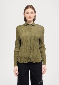 Olive green, long-sleeve button-up shirt with crinkled texture, a collared neck, and a slightly gathered hem. Paired with black cargo pants.