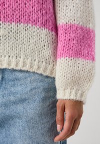 Pull tricoté à rayures roses et blanches, avec une coupe ample, des poignets côtelés, et un mélange de laine doux et texturé. Jean en denim visible en dessous.