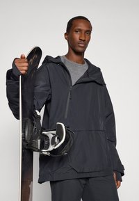 Chaqueta negra impermeable con capucha, cremallera frontal y un gran bolsillo. La persona sostiene una tabla de snowboard con fijaciones. Camiseta gris debajo.