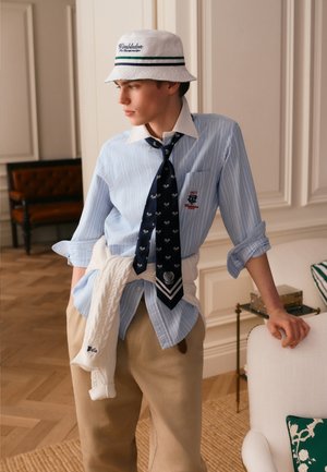 Jonge man met een Wimbledon bucket hat, blauw gestreept overhemd met marineblauwe stropdas, beige broek en witte trui gestrikt rond de taille, leunend tegen een stoel.