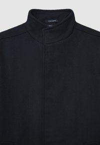 Cappotto nero con colletto alto, realizzato in tessuto testurizzato. Presenta un design minimalista con cuciture nascoste e senza hardware visibile.