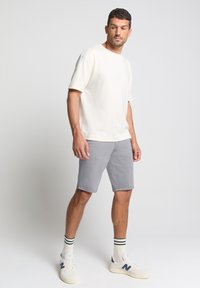 Crème katoenen T-shirt, grijze katoenen shorts en witte sneakers met marineblauwe accenten. Model staat rechtop met de handen langs het lichaam, en toont de kleding.