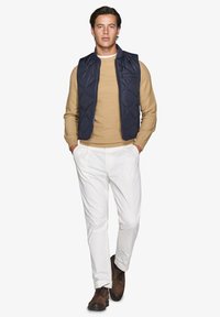 Marineblauer gesteppter Westen über einem tanfarbenen Pullover, kombiniert mit weißen Hosen und braunen Schuhen. Slim Fit; strukturierte Materialien und ein Reißverschluss.