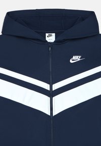 Navy modrá Nike mikina s plným zipsom, biele chevron pruhy a logo na hrudi. Ľahký materiál s textúrovaným povrchom.