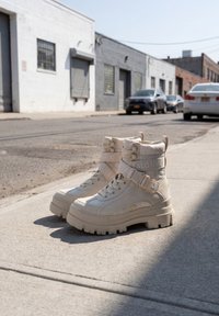 Chunky beige lace-up boots with thick soles and branded straps sit on a sunlit urban sidewalk with parked cars and buildings in the background.