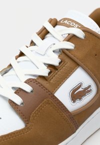 White sneaker with brown suede accents and a textured leather overlay. Features white laces and a small embroidered crocodile logo.