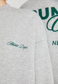 Šedá mikina s bavlny se zeleným výšivkovým textem "Athletic Dept." a tučným nápisem na zádech. Měkká textura, kulatý výstřih.