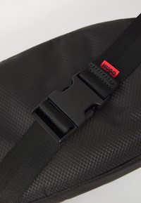Schwarzes verstellbares Band mit Plastikverschluss und kleinem rotem Etikett mit "HUGO"-Aufschrift auf strukturiertem schwarzen Stoffhintergrund.