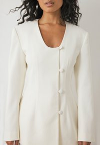 Valkoinen räätälöity bleiseri, jossa on syvä V-pääntie, pitkät hihat ja kuusi pyöreää nappisuljenta. Liukas kangas, jossa on pystysaumayksityiskohtia.