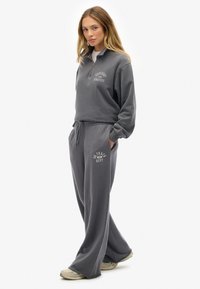 Frau steht in entspannter grauer Sportbekleidung mit einem Reißverschluss-Sweatshirt und lockeren Jogginghosen, die Hände in den Taschen, trägt beige Sneaker.
