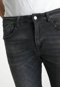 Jean en denim gris foncé avec une texture lisse, cinq poches et une fermeture à bouton argenté à la taille. Design à coupe droite.