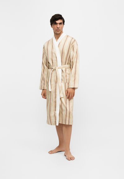 Homme debout pieds nus portant un peignoir beige à rayures jusqu’aux genoux, noué à la taille, sur un fond blanc uni.