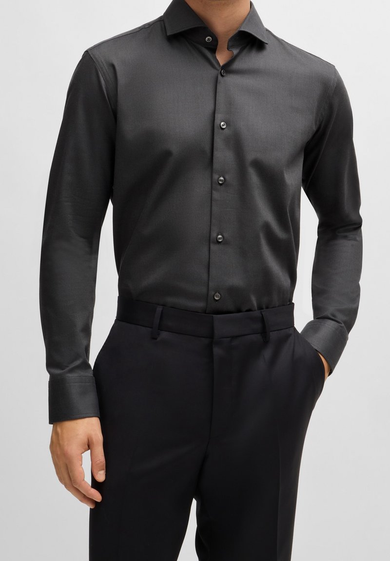 Camicia da uomo grigio scuro in tessuto liscio, con vestibilità slim, maniche lunghe, colletto appuntito e bottoni neri; abbinata a pantaloni su misura.