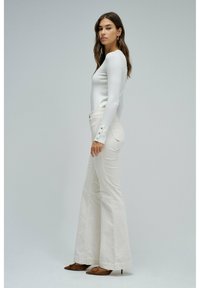 Blusa blanca de manga larga con puños abotonados combinada con pantalones de crema acampanados y zapatos marrones de punta. Diseño simple, textura suave.
