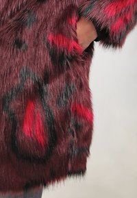 Abrigo de piel sintética en burdeos oscuro con patrones abstractos en rojo y negro. Presenta una textura suave y esponjosa y un diseño con abertura frontal.