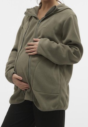 Oliwkowa polarowa kurtka zapinana na zamek z kapturem i dwoma bocznymi kieszeniami, zaprojektowana z myślą o brzuszku ciążowym. Miękka w dotyku, luźny krój.