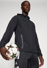 Zwarte sportieve jas met capuchon, ritszakken en grijze accenten. De drager houdt een patroonvoetbal vast, gecombineerd met een zwarte broek.