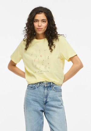Femme aux cheveux bouclés portant un t-shirt jaune avec un motif floral et le texte "Le Jardin", associé à un jean bleu taille haute, les mains sur les hanches.