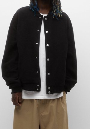 Personne portant une veste noire boutonnée sur une chemise blanche et un pantalon beige, debout devant un fond clair uni.