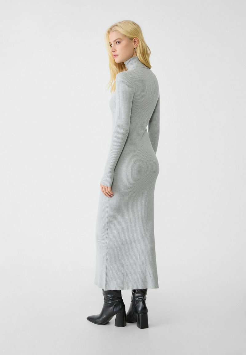 Vestido de cuello alto de manga larga y canalé en gris claro, con silueta recta, aberturas laterales, combinado con botas negras de tacón.